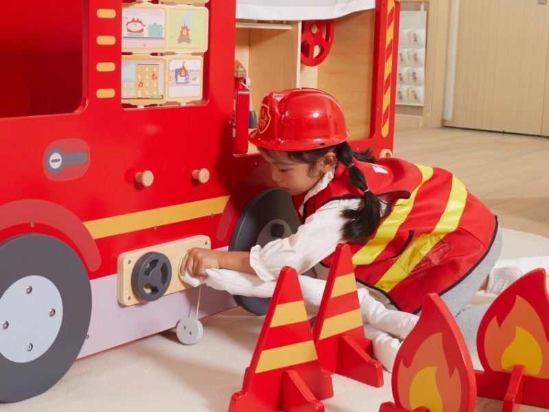 Mädchen in Feuerwehr-Kleidung schliesst einen Schlauch an ein Spielmöbel-Feuerwehrauto an