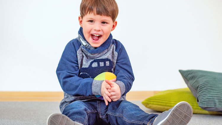 Junge sitzt lachend auf dem Boden und hält einen Ball aus dem Betzold Online-Shop in der Hand.